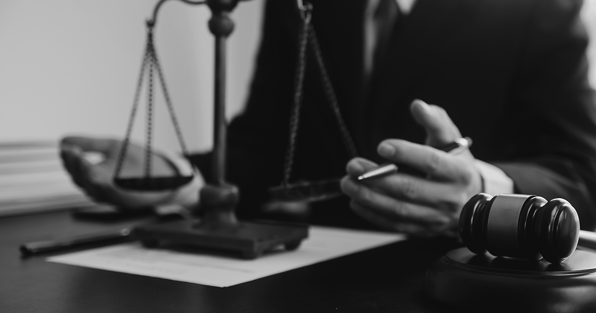 Image of an unseen person with a pen in their hand sat behind a set of scales and a gavil