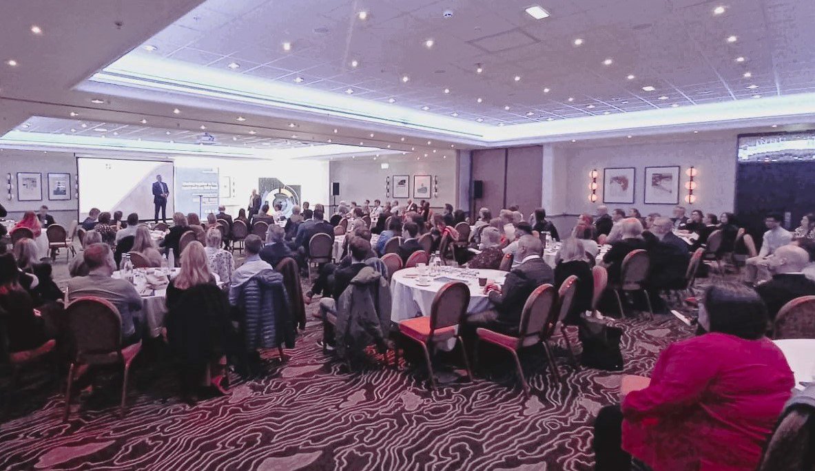 Image of room full of people, sitting listening to a speaker on stage