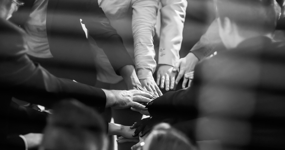 Image showing a large group of people putting their hands in the middle of a circle to symbolise working together.