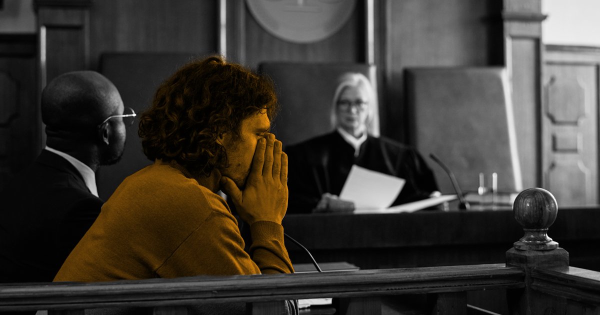 Image of a person sitting with their face in the hands, with a judge looking at them reading from a document infront of them.